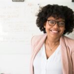 A smiling woman wearing glasses and a blush blazer stands in front of a whiteboard filled with red marker notes, conveying confidence and professionalism.